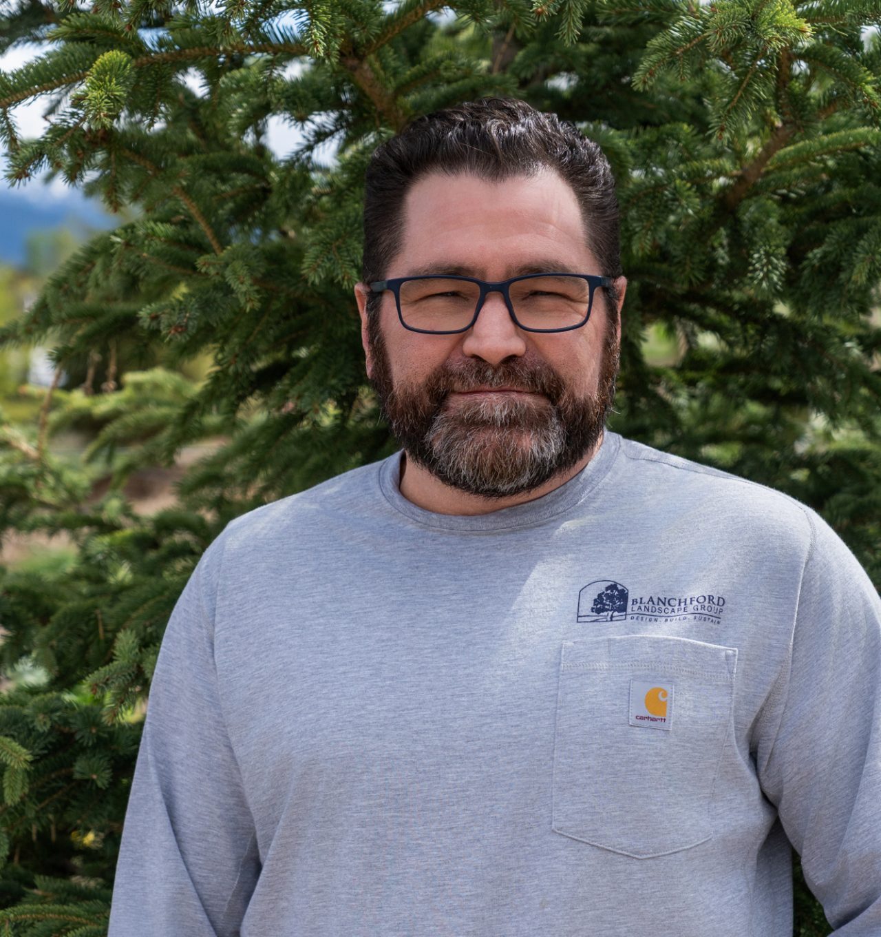 Bearded man in glasses standing before evergreen trees